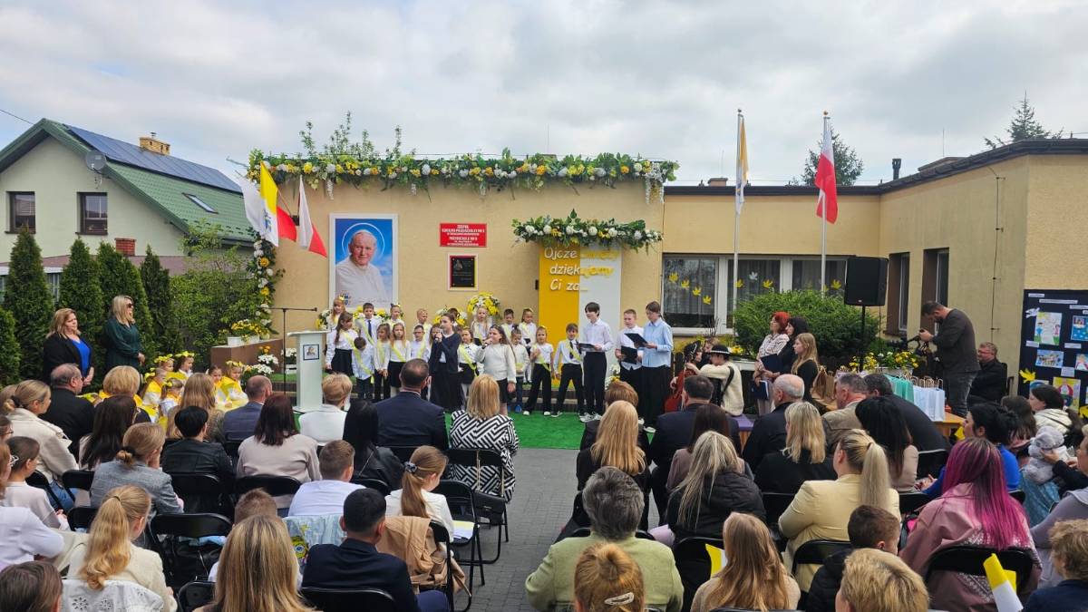 Na zdjęciu Jubileusz 20-lecia nadania imienia Jana Pawła II Przedszkolu nr 9. Uczestnicy uroczystości siedzą na placu przedszkolnym, wkoło barwy papieskie i chorągiewki