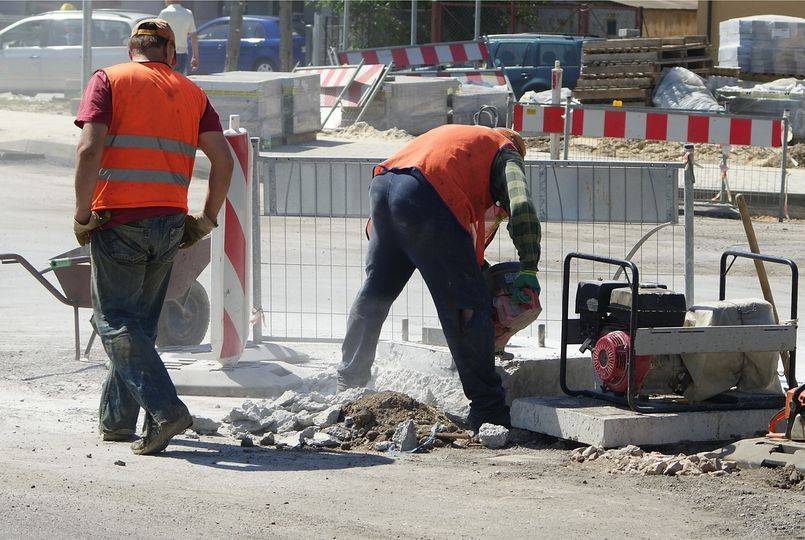 Na zdjęciu widać dwóch pracowników drogowych podczas remontu ulicy. Obaj mają na sobie pomarańczowe kamizelki odblaskowe i robocze ubrania. Jeden z nich rozbija nawierzchnię drogi, a drugi stoi obok z taczką. Wokół znajdują się barierki zabezpieczające.