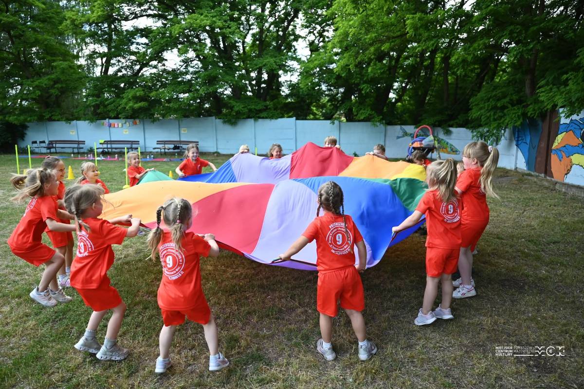 Na zdjęciu dzieci tańczą trzymając wielokolorową płachtę na terenie zielonym MCK Za Pilicą