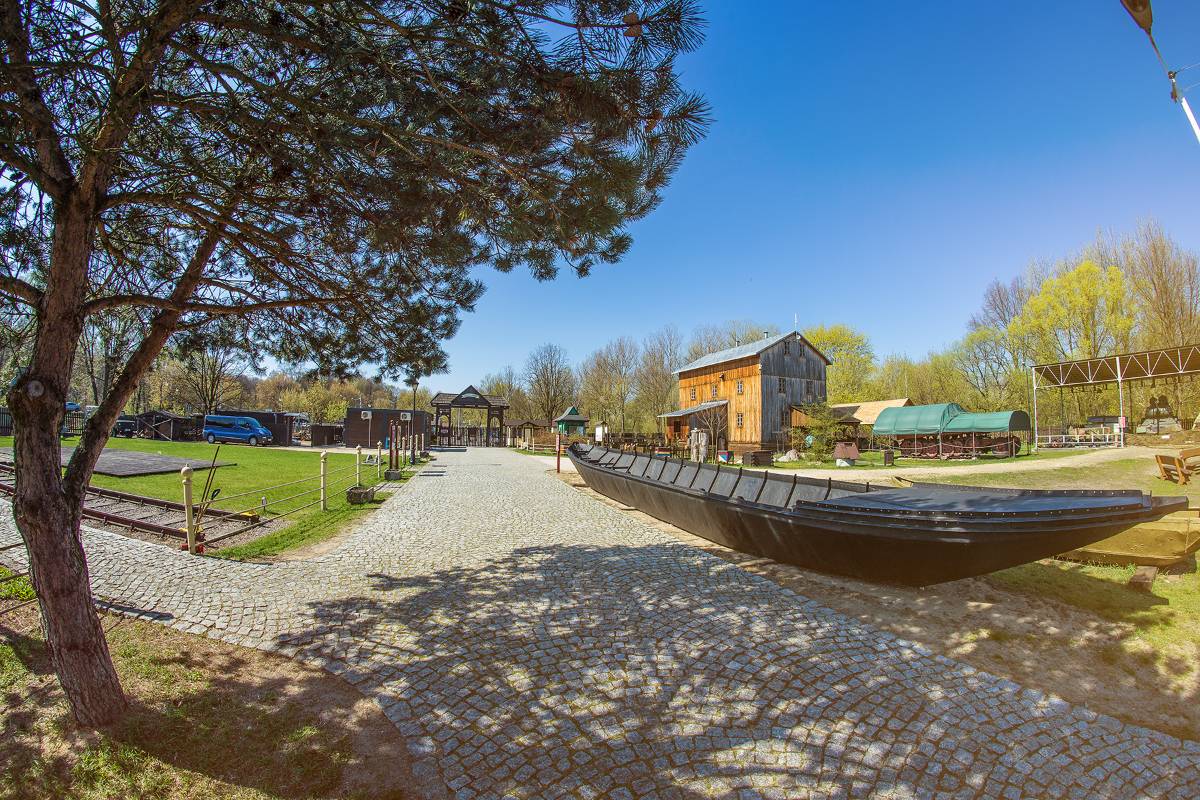 Na zdjęciu Skansen rzeki Pilicy, na pierwszym planie widać łódź Szczupak, na dalszym planie drewniany budynek dawnego młyna. Widać również ścieżkę wyłożoną drobną kostką brukową i drzewo