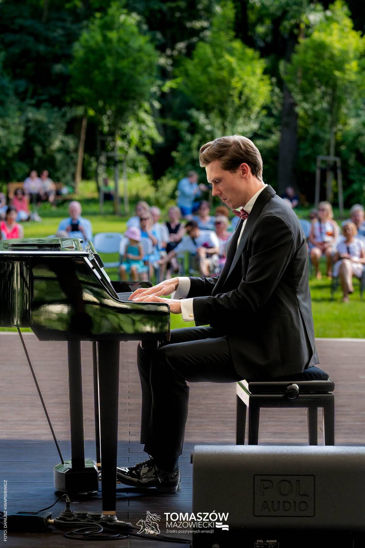 Na zdjęciu pianista przy fortepianie gra utwory Chopina. W tle publiczność zgromadzona w Parku Solidarność. Pianista w garniturze, białej koszuli z czerwoną muchą.