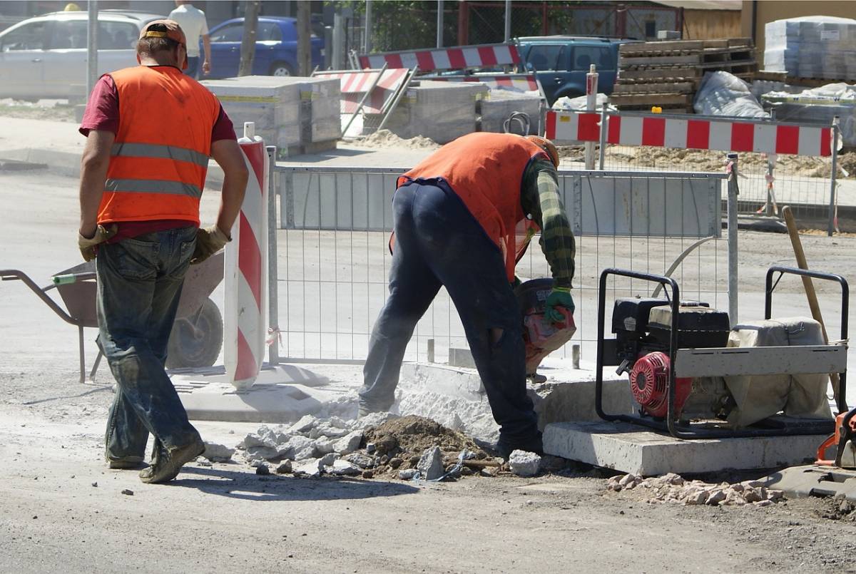 Robotnicy w pomarańczowych kamizelkach podczas robót drogowych. W tle narzędzia m.in. taczka, barierki zabezpieczające, betonowe płyty, gruz.
