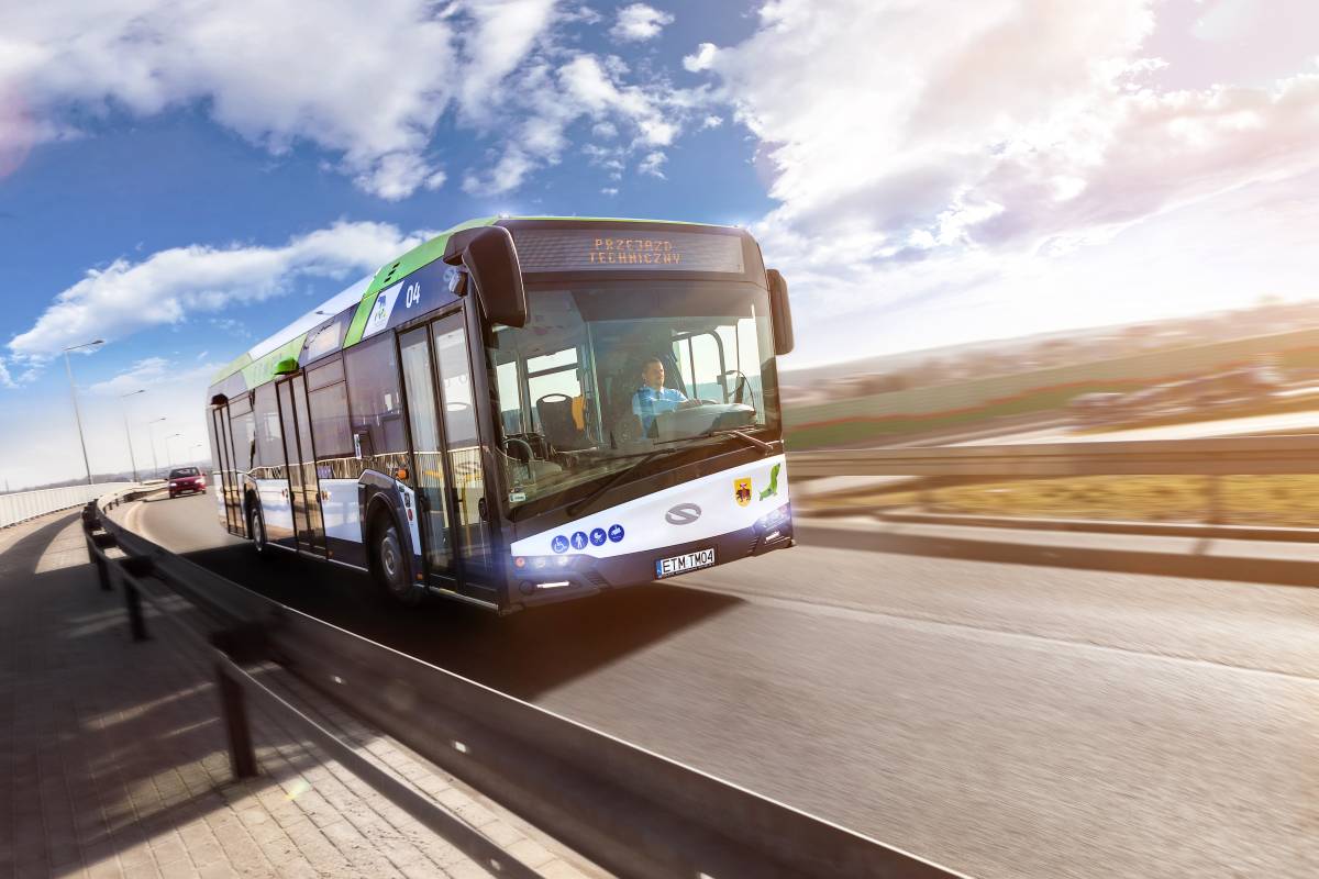 Na zdjęciu autobus MZK przejeżdżający przez tamę Zalewu Sulejowskiego, przez przednią szybę widać kierowcę, w tle zbiornik wodny i pola