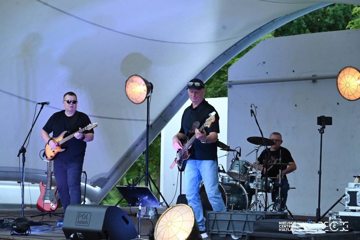 Na zdjęciu trio rockowe Kazzu podczas występu w ramach Letniej Sceny Artystycznej w amfiteatrze Przystani nad Pilicą. Z przody dwóch gitarzystów z tyłu perkusista, wszyscy w czarnych t-shirtach