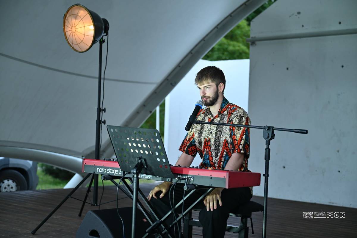 Max Miszczyk ubrany w koszulę z etrycznym wzorem, siedzi przy czerwonym pianinie scenicznym i śpiewa do mikrofonu.