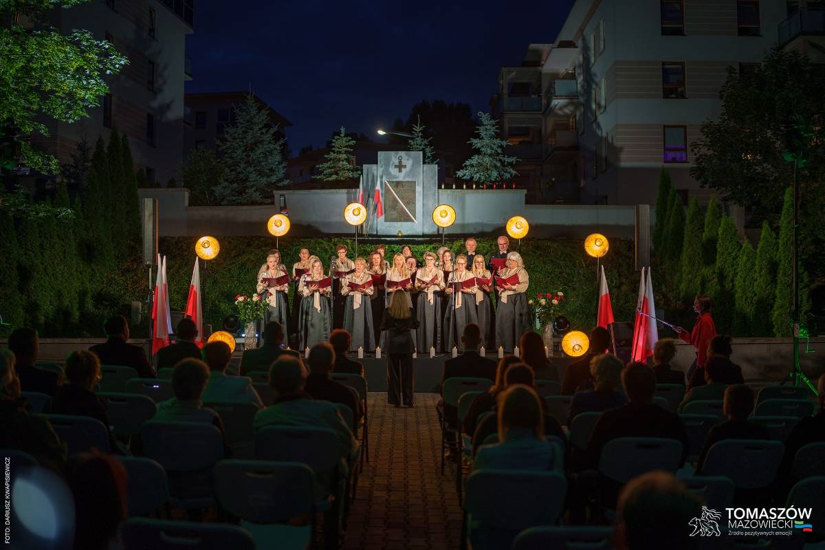 Koncert Chóru Artis Gaudium przed Pomnikiem Ku Czci Rozstrzelanych Członków Ruchu Oporu i Zakładników w latach 1939–1945 przy ul. Browarnej. Przed chórzystami dyrygent oraz zgromadzona publiczność. Koncert odbywa się po zmroku, w otoczeniu zieleni.