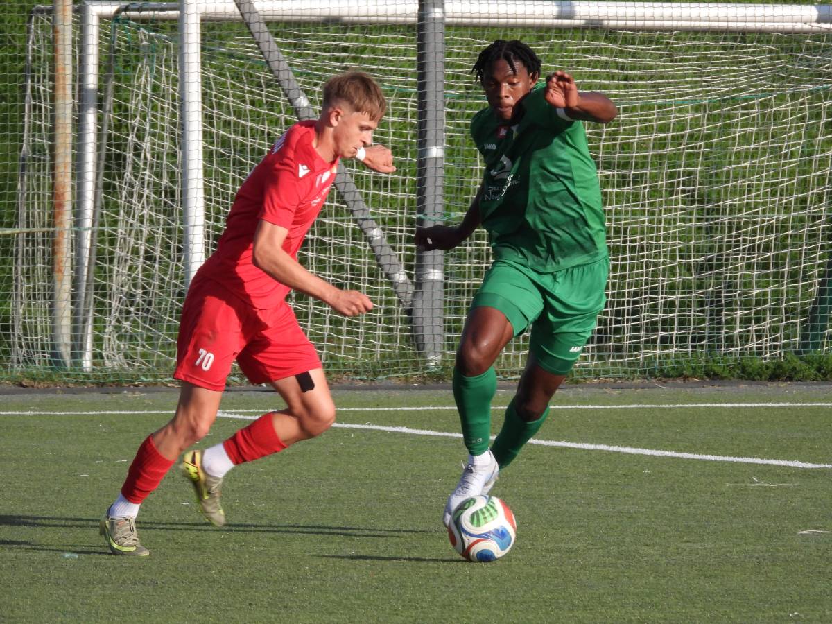 Mecz Lechia -Świt rozgrywany w słoneczny dzień na boisku z zieloną, sztuczną murawą. W tle widoczna metalowa konstrukcja bramki oraz siatka. Przy piłce zawodnik w zielonym stroju próbuje ominąć przeciwnika. Piłkarz Lechii w czerwonym stroju walczy o piłkę