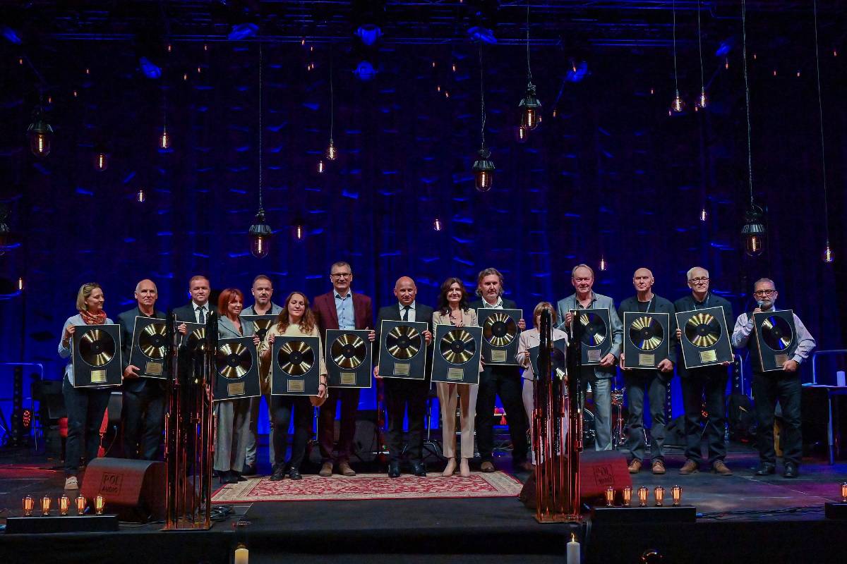 Grupa kilkunastu osób stoi na zaciemnionej scenie z profesjonalym, niebieskim oświetleniem w tle. Zgromadzeni trzymają Płote Płyty, które otrzymali za współtworzenie Festiwalu i wkład w rozwój polskiego jazzu