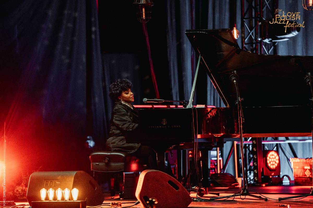 Kobieta gra na fortepianie podczas Love Polish Jazz Festival. Artystka siedzi profilem do widza i śpiewa do mikrofonu. Scena jest oświetlona ciepłym pomarańczowo-czerwonym światłem. W dolnej części widoczny zarys publiczności festiwalowej