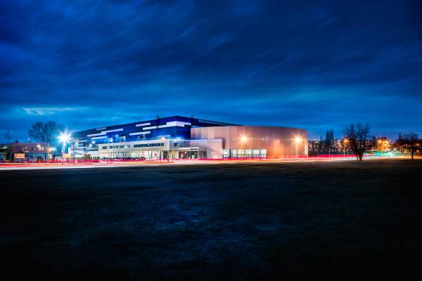 Nocny widok na budynek Areny Lodowej w Tomaszowie Mazowieckim, oświetlony niebieskimi i białymi światłami, z widocznymi smugami świateł przejeżdżających samochodów.