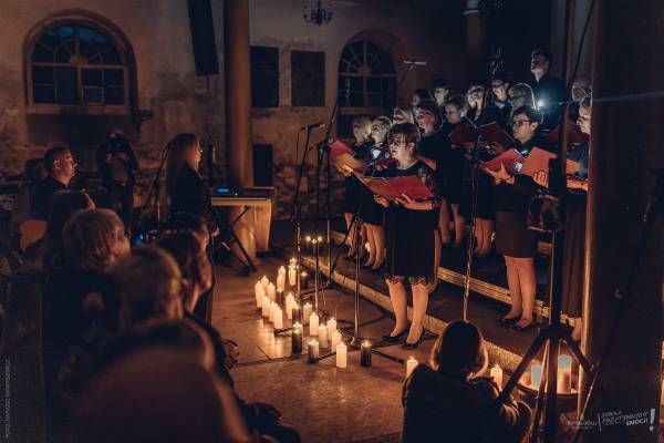 Występ chóru w zabytkowym wnętrzu z kolumnami, oświetlonym rzędem świec ustawionych na podłodze; widok z perspektywy siedzącej publiczności.