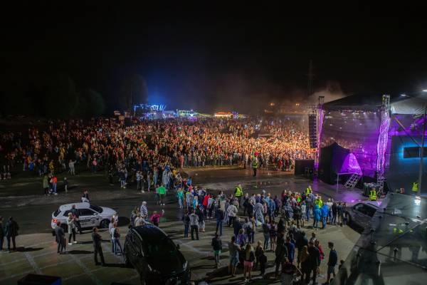 Nocny koncert plenerowy z tłumem licznie zgromadzonej publiczności przed sceną oświetloną fioletowymi światłami, w tle stoiska festiwalowe i samochody.