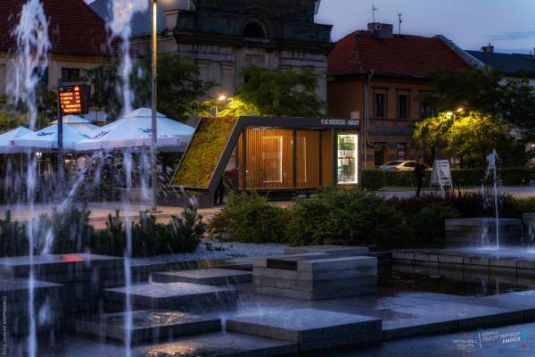 Wieczorny widok na plac miejski z tryskającą fontanną, nowoczesnym pawilonem informacyjnym z zielonym dachem oraz zabytkowymi kamienicami w tle.