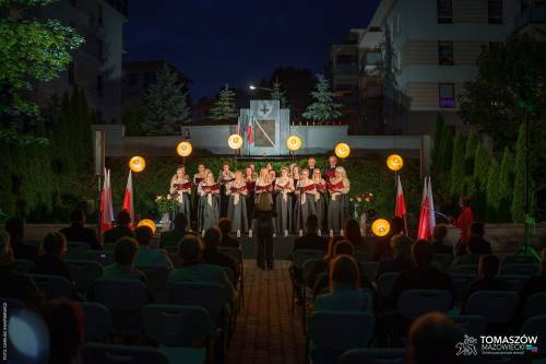 Koncert Chóru Artis Gaudium przed Pomnikiem Ku Czci Rozstrzelanych Członków Ruchu Oporu i Zakładników w latach 1939–1945 przy ul. Browarnej. Przed chórzystami dyrygent oraz zgromadzona publiczność. Koncert odbywa się po zmroku, w otoczeniu zieleni.