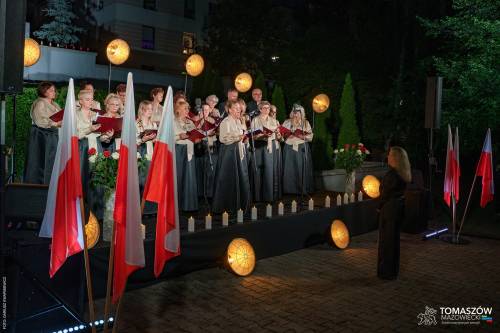 Koncert Chóru Artis Gaudium przed Pomnikiem Ku Czci Rozstrzelanych Członków Ruchu Oporu i Zakładników w latach 1939–1945 przy ul. Browarnej. Chórzyści w jednakowych strojach,przed nimi dyrygent. Koncert odbywa się po zmroku, w otoczeniu zieleni.