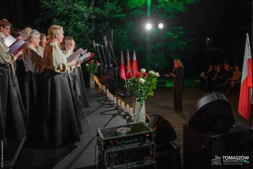 Koncert Chóru Artis Gaudium przed Pomnikiem. Wykonawcy w otoczeniu świateł i biało-czerwonych flag Polski. Przed nimi dyrygent i publiczność. Koncert odbywa się po zmroku, w otoczeniu zieleni.