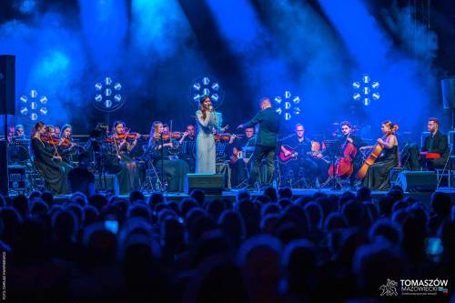 Koncert Katarzyny Zawady z TM Orchestra. Na środku sceny stoi wokalistka, ubrana w długą, białą suknię i śpiewa do mikrofonu. Otacza ją orkiestra. Muzycy siedzą i grają m.in. na skrzypcach i wiolonczeli. Dyrygent stoi tyłem do publiczności