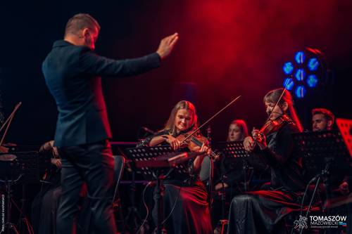 Koncert Katarzyny Zawady z TM Orchestra. N scenie muzycy Tomaszowskiej Orkiestry Symfonicznej. Graja m.in. na skrzypcach, wiolonczelach. Kobiety ubrane są w ciemne, wieczorowe suknie. Przed muzykami stoi dyrygent z podniesionymi rękami