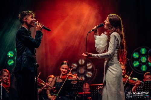 Koncert Katarzyny Zawady z TM Orchestra. N scenie duet - mężczyzna i kobieta śpiewają do mikrofonów. Wokalistka ma na sobie długą, białą suknię. Mężczyzna jest ubrany w czarny garnitur. Za nimi siedzą i grają muzycy Tomaszowskiej Orkiestry Symfonicznej