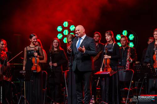 Koncert Katarzyny Zawady z TM Orchestra. N scenie Prezydent Tomaszowa Mazowieckiego w otoczeniu stojących muzyków Tomaszowskiej Orkiestry Symfonicznej. Prezydent z uśmiechem przemawia do publiczności. Scena podświetlona na czerwono.