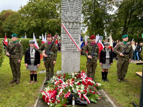 W centrum Pomnik „W Hołdzie Zesłańcom Sybiru”. Przy pomniku warta honorowa harcerzy i wojska. Obelisk zdobi biało-czerwona wstęga i złożone prze delegacje kwiaty. W tle uczestnicy z flagami narodowymi na drzewcach.