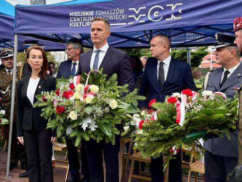 Uczestnicy obchodów patroiotycznych stoją pod namiotem Miejskiego Centrum Kultury. Wsród zgromadzonych znajdują się m.in.  przedstawiciele władz miejskich i powiatowych, służb mundurowych i wojska. Niektórzy trzymają wieńce biało-czerwonych kwiatów