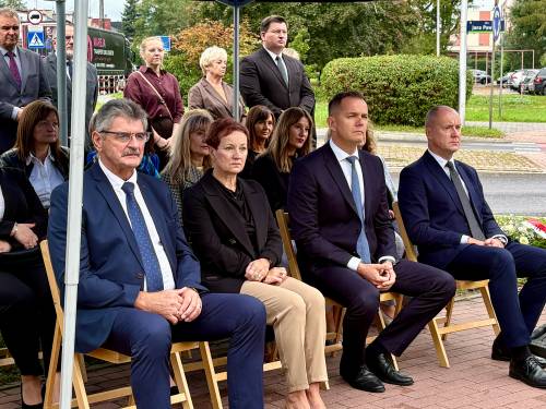 Uczestnicy uroczystości patriotycznej siedzą na krzesłach przed pomnikiem  „W Hołdzie Zesłańcom Sybiru”. Wśród nich znajdują się m.in. przedstawiciel władz miejskich. Wszyscy są ubrani w eleganckie stroje. W tle widoczna jest infrastruktua miejska