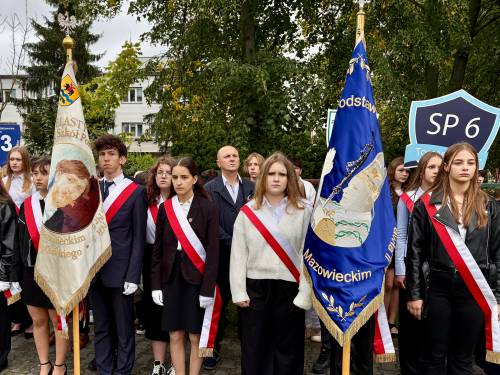 Poczty sztandarowe przed kościełem pw. NMP Królowej Polski przy ul. Słowackiego. Na pierwszym planie uczniowie w galowych strojach i biało-czerwonych szarfach. Część z nich trzyma na drzewcach sztandary. W tle widoczne tablice tarcz szkolnych m.in. SP 6