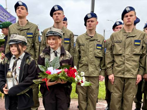 Młodzi mężczyźni ze szkoły mundurowej  stoją przed Pomnikiem „W Hołdzie Zesłańcom Sybiru”.  Przed nimi stoją harcerki. Jedna z nich trzyma bukiet biało-czerwonych kwiatów, przewiąznych biało-czerwoną wstęgą, a druga znicz