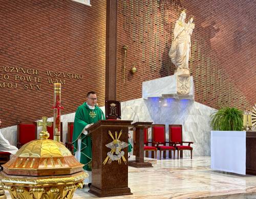 Przy ambonie kapłan w zielonej szacie liturgicznej odprawia mszę św. w kościele  pw. NMP Królowej Polski przy ul. Słowackiego. W tle wysoka ściana z czerwonej cegły i figura Matki Boskiej. Po lewj stronie, przed kapłanem stoi złocona chrzcielnica