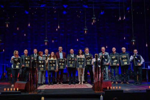 Grupa kilkunastu osób stoi na zaciemnionej scenie z profesjonalym, niebieskim oświetleniem w tle. Zgromadzeni trzymają Płote Płyty, które otrzymali za współtworzenie Festiwalu i wkład w rozwój polskiego jazzu