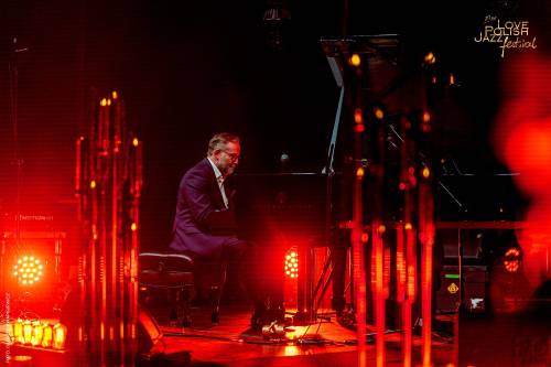 Mężczyzna w garniturze gra na fortepianie podczas Love Polish Jazz Festival. Muzyk siedzi profilem do widza i jest pochylony nad instrumentem. Scena jest oświetlona ciepłym pomarańczowo-czerwonym światłem.