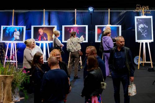 Grupa ludzi zgromadzona w zaciemnionej części Areny Lodowej. W tle na sztalugach stoją fotografie (autorstwa Dariusza Kwapisiewicza) z artystami z  poprzednich edycji festiwalu. Uczestnicy wydarzenia oglądają wystawę i rozmaiwają.