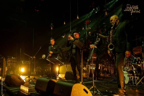 Zespół muzyczny na zaciemnionej scenie gra koncert w czasie festiwalu jezzowego. W centralnej części artyści z instrumentami m.in. saksofonista. Na podłodze stoją wlwmwnty nagłośnienia i reflektory - emitujące klimatyczne żółto-pomarańczowe światło