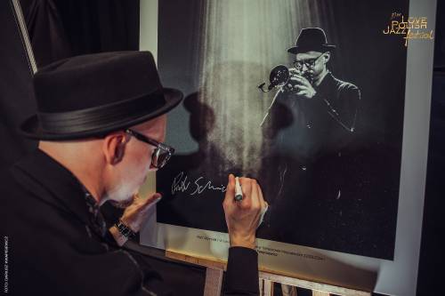 Mężczyzna w okularachi kapeluszu trzyma w dłoni czarny marker, którym podpisuje swój duży, czarno-biały portret. Zdjęcie przedstawia trębacza i pochodzi z poprzedniej edycji festiwalu.