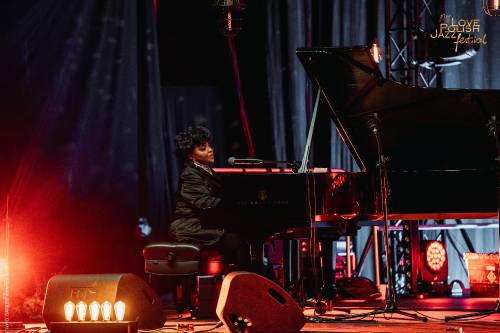 Kobieta gra na fortepianie podczas Love Polish Jazz Festival. Artystka siedzi profilem do widza i śpiewa do mikrofonu. Scena jest oświetlona ciepłym pomarańczowo-czerwonym światłem. W dolnej części widoczny zarys publiczności festiwalowej
