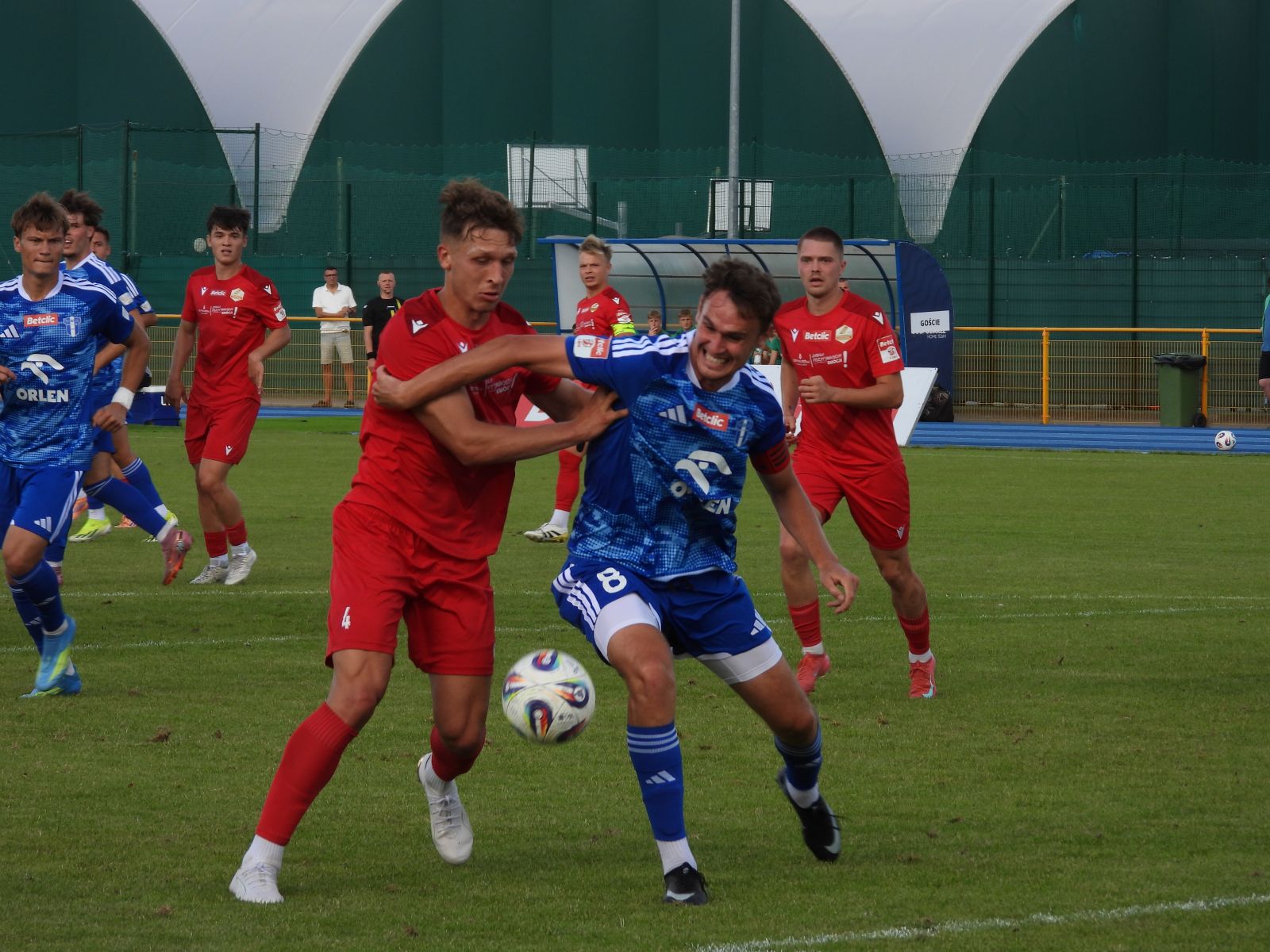 Zawodnik Wisły Płock II (w niebieskim stroju walczy) o piłkę z piłkarzem Lechii (w czerwonym). Obaj zawodnicy są w biegu, piłakrz Wisły II Płock próbuje zatrzymać przeciwnika, obejmując go ramieniem. W tle widoczni są pozostali zawodnicy obu drużyn oraz ławka rezerwowych.