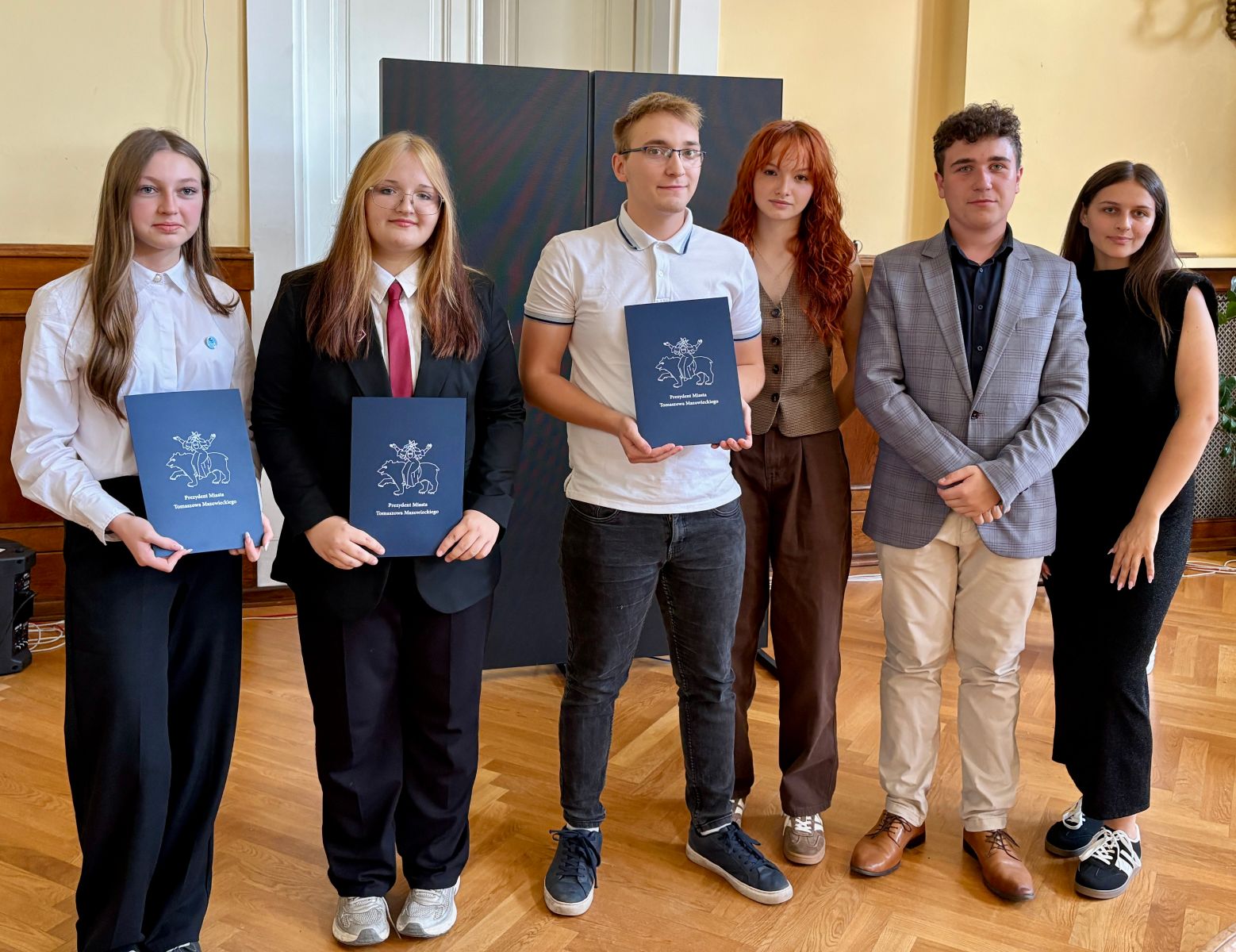 Pamiątkowe zdjęcie przedstawicieli Młodziezowej Rady Miasta (trzy kobiety i dwóch mężczyzn) . Od lewej  trójka Radnych, kończących swoja pracę na rzecz miasta. W rękach trzymają granatowe teczki z logo Tomaszowa Mazowieckiego.