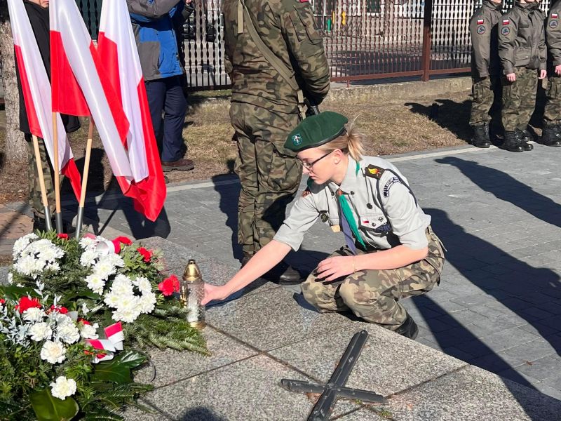 Na zdjęciu kwiaty pod tablicą płk. Witolda Pileckiego. Obok biało-czerwone flagi na stojakach