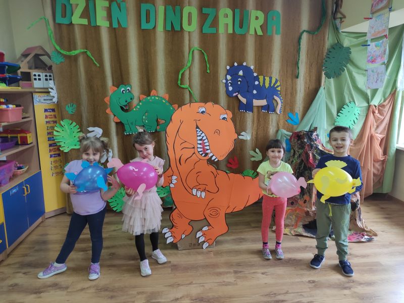 Na zdjęciu przedszkolaki w trakcie Tygodnia z Dinozaurami. Na zdjęciu kartonowy dinozaur i dzoieci z dinozauramiz balonów